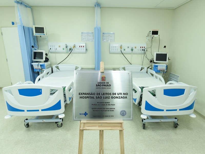 Foto de dois dos dez leitos entregues no Hospital São Luiz Gonzaga. À frente, em primeiro plano, está a placa de inauguração com o emblema da cidade de São Paulo. Dois leitos aparecem ao fundo. Eles são brancos com detalhes em azul e cercados por aparelhos. Do teto sai uma cortina azul que separa os dois leitos
